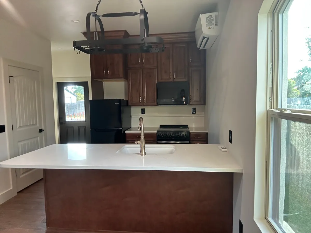 White peninsula countertop with a sink, looking toward the wood kitchen cabinets, a black refrigerator, and the back door, with a pot rack above.