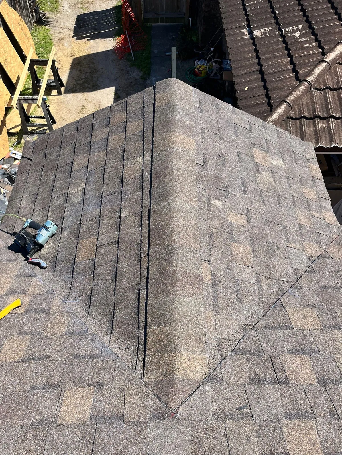 Close view of shingled roof planes meeting at a valley on each side of a ridge, with a nail gun resting on the roof.
