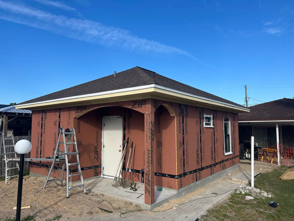Corner view of the sheathed studio with its arched porch, a white door, and ladders, before stucco.