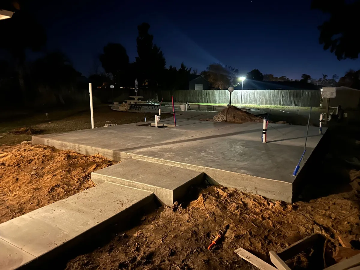 Concrete slab and adjoining porch pad under work lights in the evening.