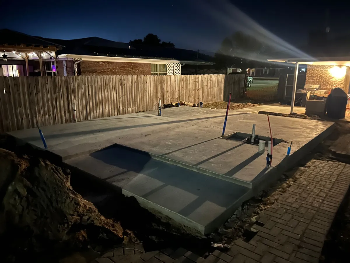 Freshly poured concrete slab at dusk, with plumbing stubs and a paver walk alongside.