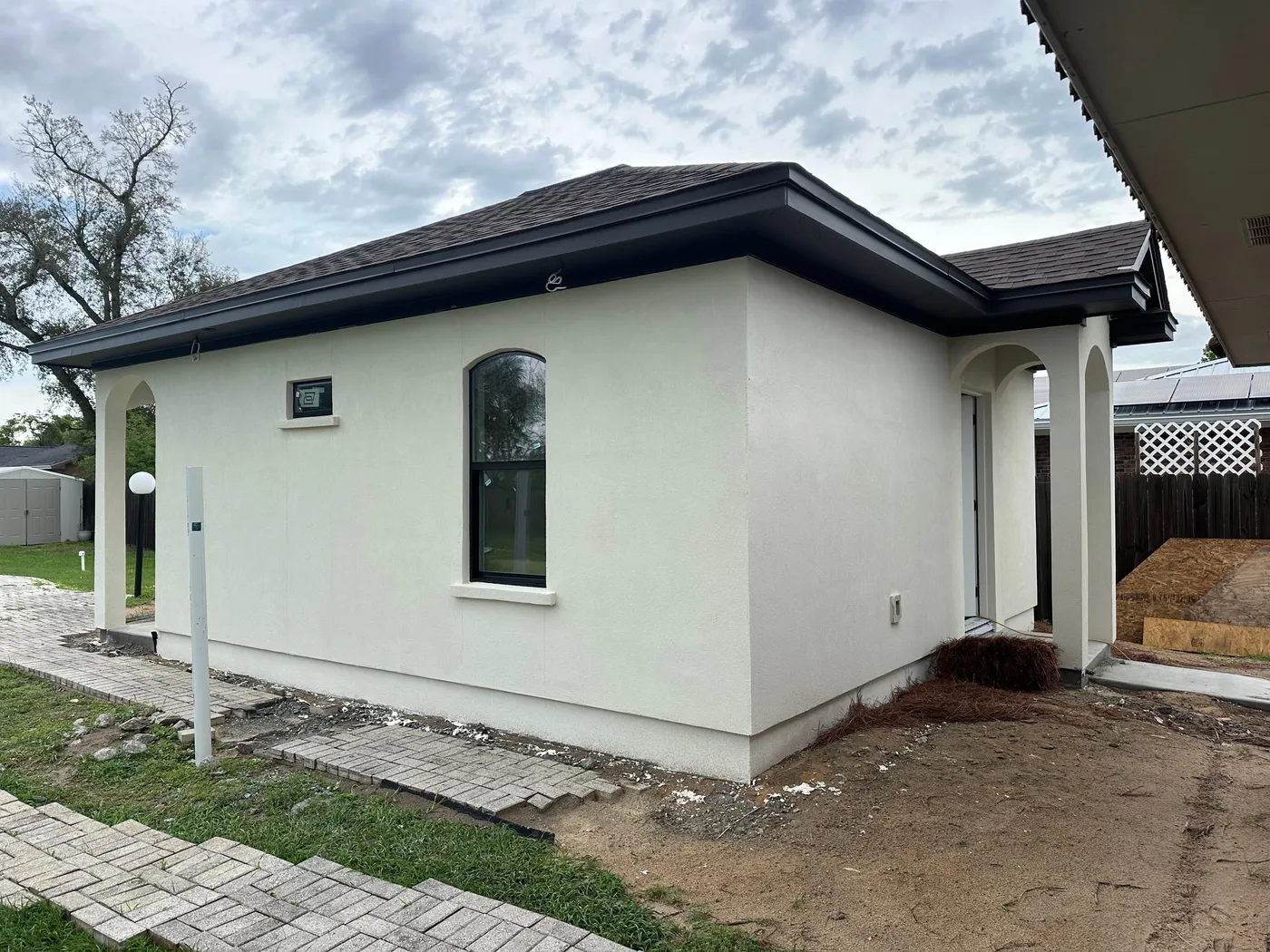 Side of the stucco studio with a recessed arched window, a small high window, and broad solid wall areas, with the arched entry at right and site work still underway.
