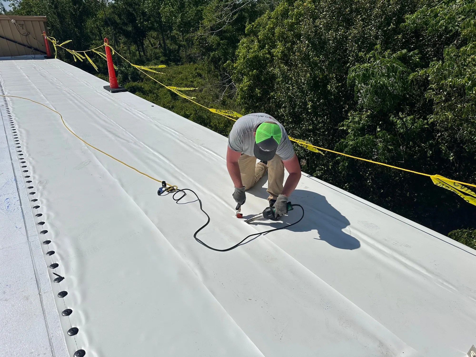 A Garrett Construction crew member hot-air welding a seam in the white membrane, with safety tape along the roof edge.