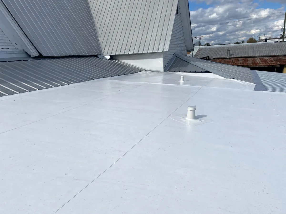 White membrane covering the former junctions below the two metal-roofed gables, with a vent penetration.