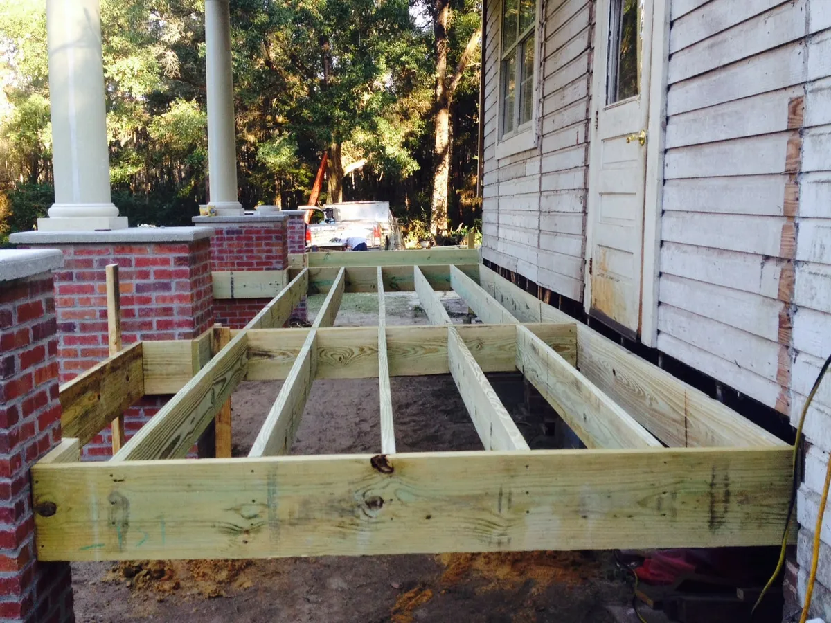New treated floor joists framed between the building wall and the brick porch piers, with a column standing beyond.