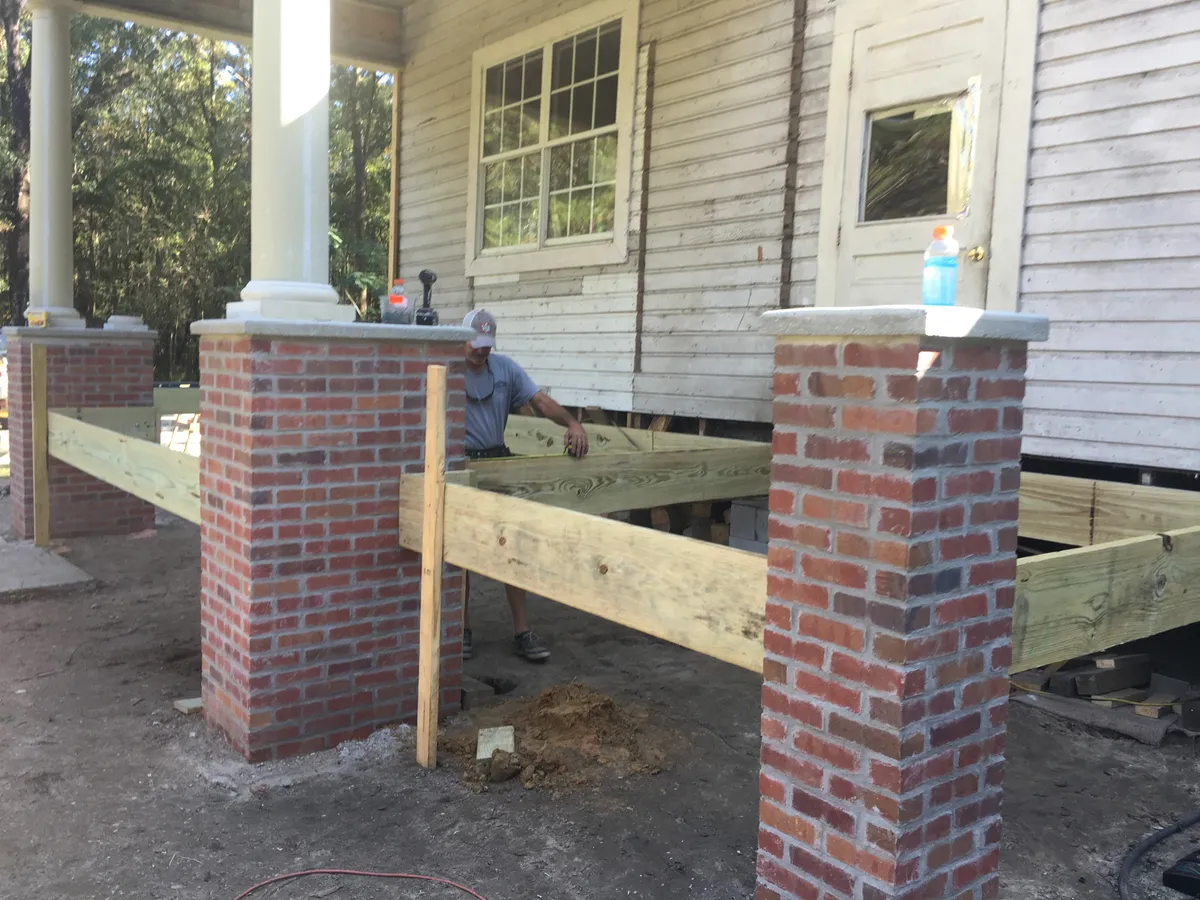 A worker setting a treated beam between capped brick piers, with a porch column and the building wall behind.